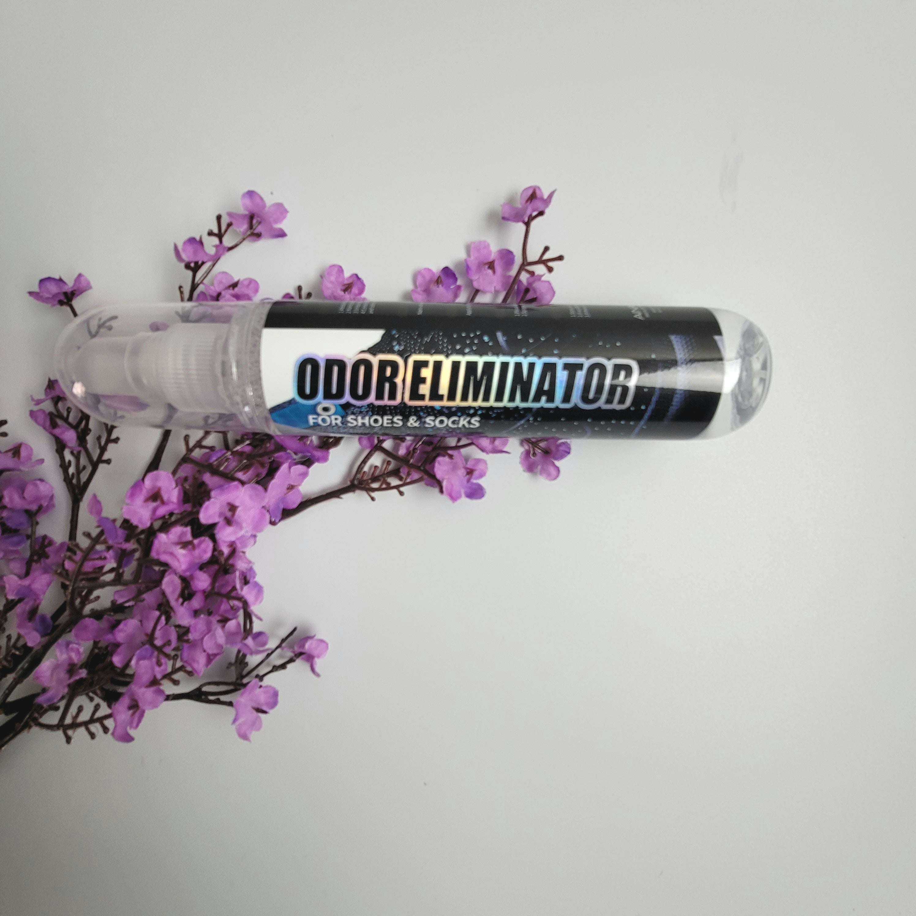 Side view of a bottle of Odor Eliminator from AinaCare laying on purple flowers on a white background.