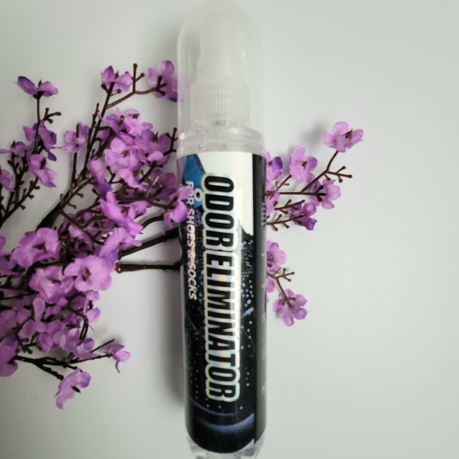 A bottle of Odor Eliminator from Aina Care on top of purple flowers on a white background.