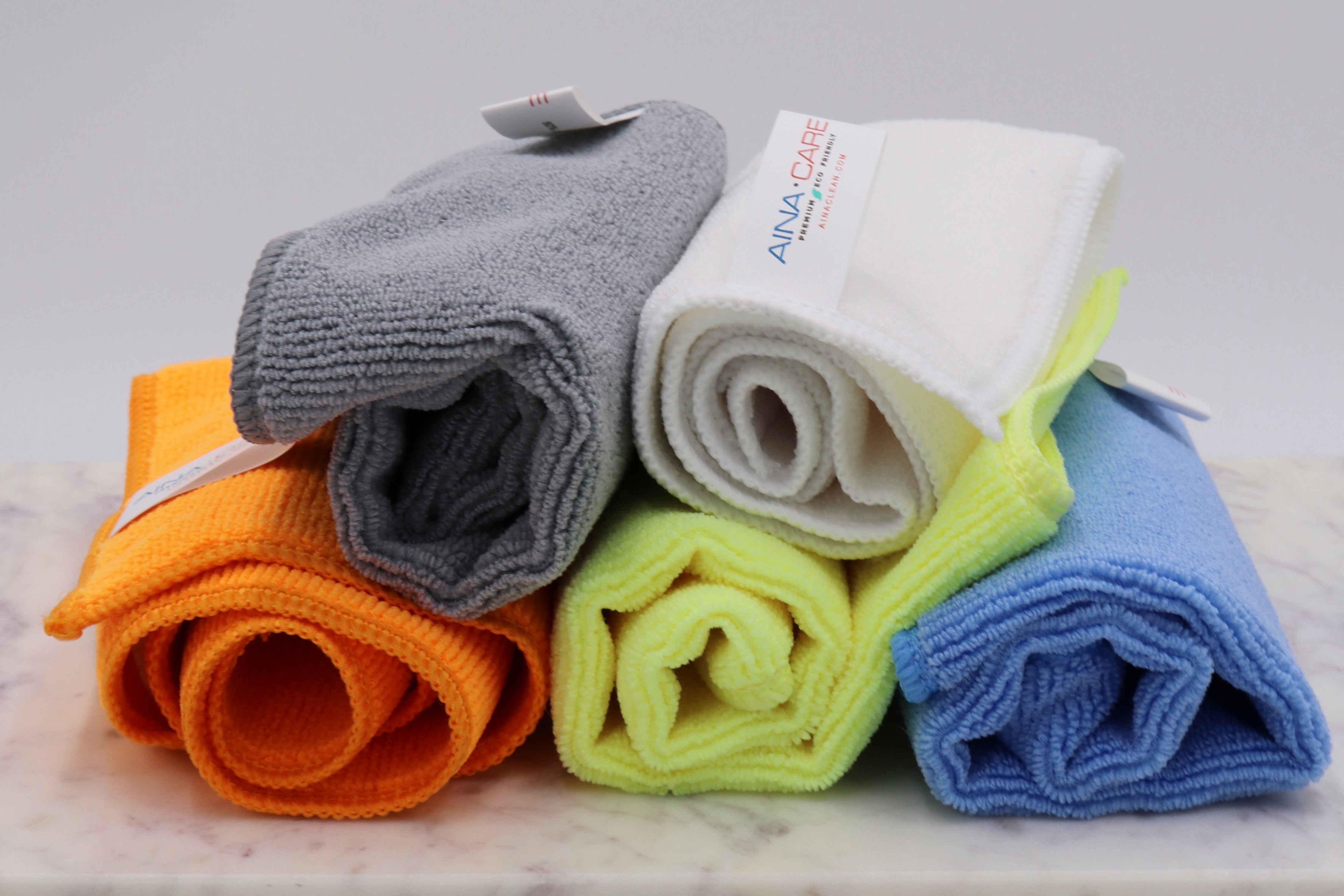 Close up view of microfiber cloths from AinaCare in each color (grey, white, orange, neon yellow, and blue).
