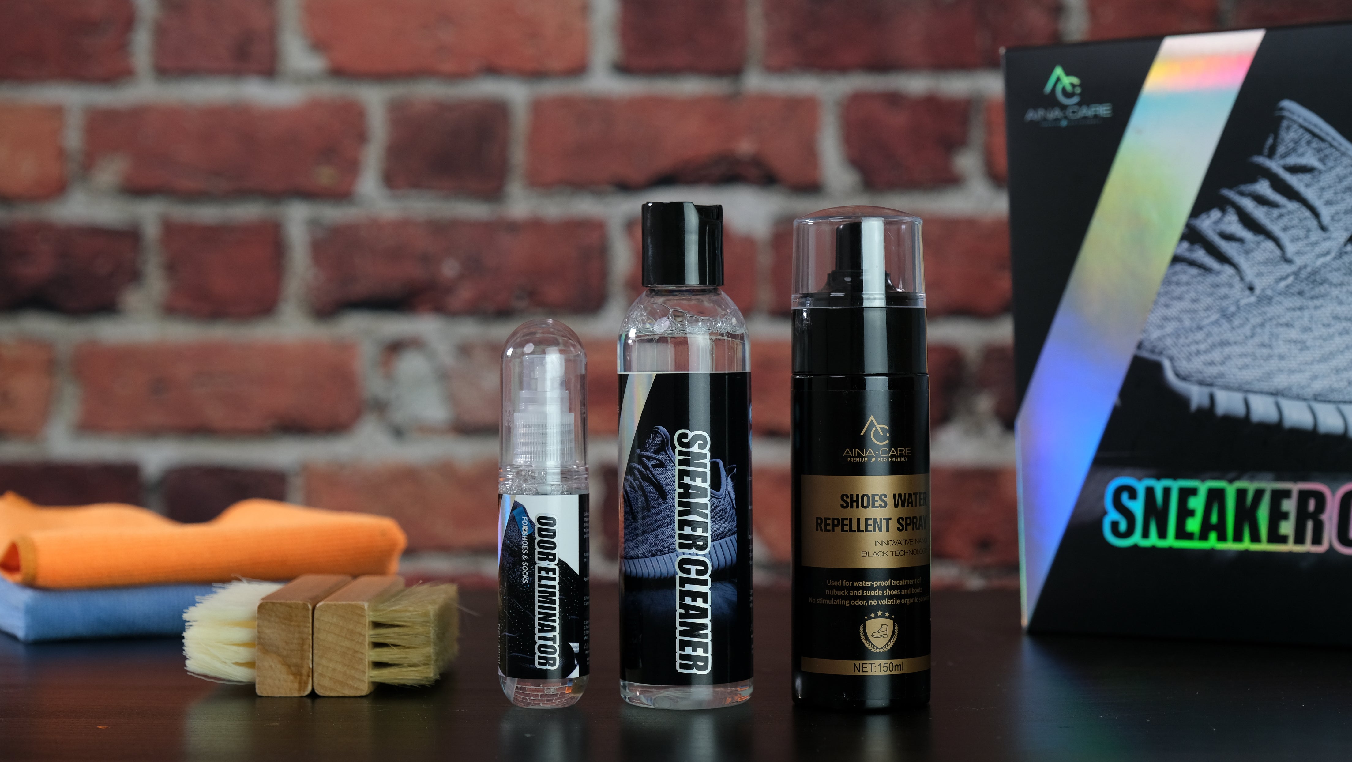 Each of the items from the Signature Premium Sneakers Kit displayed on a black table in front of a brick wall. Items from left to right are: two microfiber cloths, two brushes, an odor eliminator spray, a bottle of sneaker cleaner, a bottle of water repellent spray, the package box..