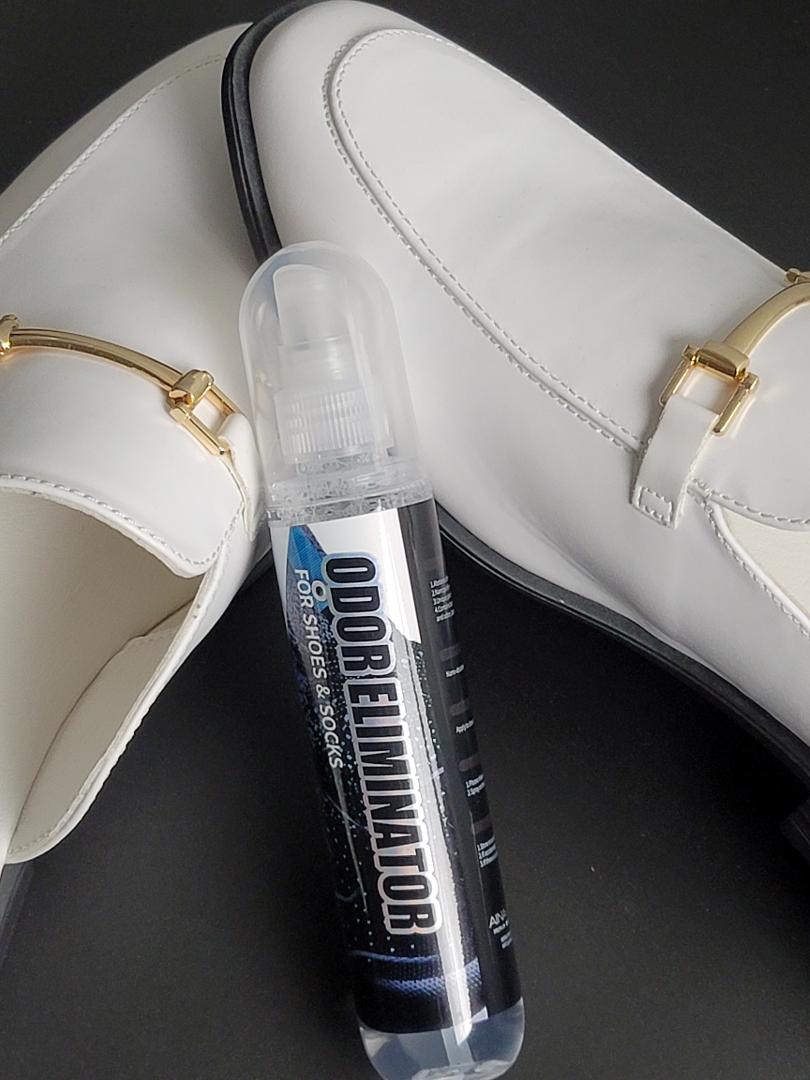 A bottle of Odor Eliminator from AinaCare propped up between two white leather loafer shoes.